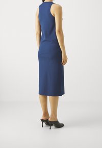 Kobieta w dopasowanej, bezrękawnikowej granatowej midi sukience i czarnych szpilkach, stojąca plecami do kamery na białym tle.
