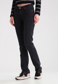 Jeans de mezclilla negros con un ajuste slim, que cuentan con un diseño de cinco bolsillos y un cierre de botón. Combinados con zapatillas negras y rosas.