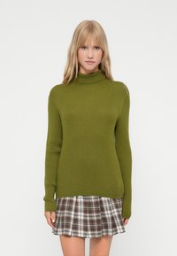 Maslinasto zelena rebrasta pletena majica s turtleneck ovratnikom i dugim rukavima, u kombinaciji s kariranom suknjom u smeškastim i belim tonovima.