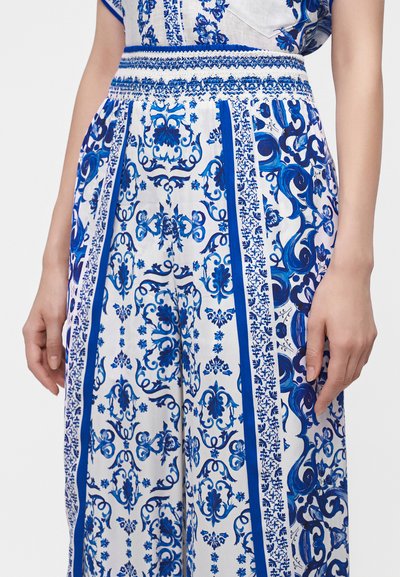 Pantalon blanc avec des motifs floraux et ornementaux bleus, doté d'une large taille smockée et de bandes décoratives verticales sur les côtés.