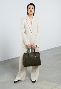 Un sac à main noir structuré avec une texture lisse et une poche zippée. Porté avec un costume à carreaux gris clair. Le modèle se tient sur un sol en béton.