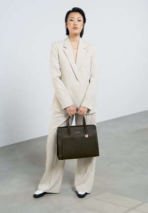 Un sac à main noir structuré avec une texture lisse et une poche zippée. Porté avec un costume à carreaux gris clair. Le modèle se tient sur un sol en béton.