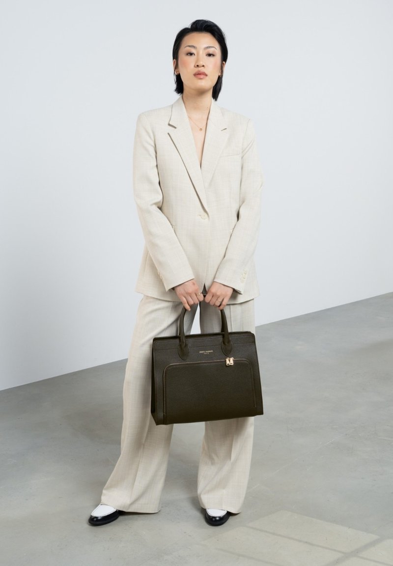 Un sac à main noir structuré avec une texture lisse et une poche zippée. Porté avec un costume à carreaux gris clair. Le modèle se tient sur un sol en béton.