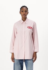 Striped long-sleeve shirt in pink and white, featuring a button-down front and a red logo patch on the left chest. Cotton fabric.