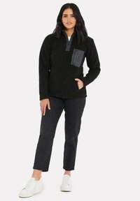 Pull en polaire noir avec un tissu texturé, col zippé et une poche sombre contrastante sur la poitrine. Associé à un jean noir et des baskets blanches.