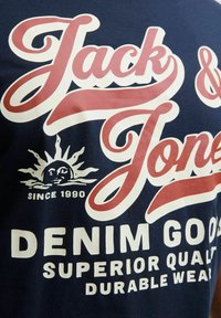 Marineblå t-shirt med hvid og rød grafisk tekst "Jack & Jones" og et sol-logo. Teksten inkluderer "DENIM GOODS", "SUPERIOR QUALITY" og "DURABLE WEAR".