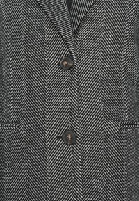 Cappotto in bianco e nero con motivo a zigzag, dotato di revers a punta, due bottoni e tasche laterali con finitura testurizzata.