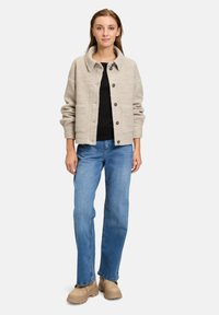 Veste beige texturée avec un col, des boutons à l'avant et deux grandes poches ; portée sur un haut noir avec un pantalon en denim bleu et des bottes beige.