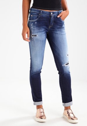 Nærbillede af en person iført falmede blå slidte jeans med opkantsøm, sort top og metalliske rosa guld slip-on sneakers, stående med hænderne i lommerne.