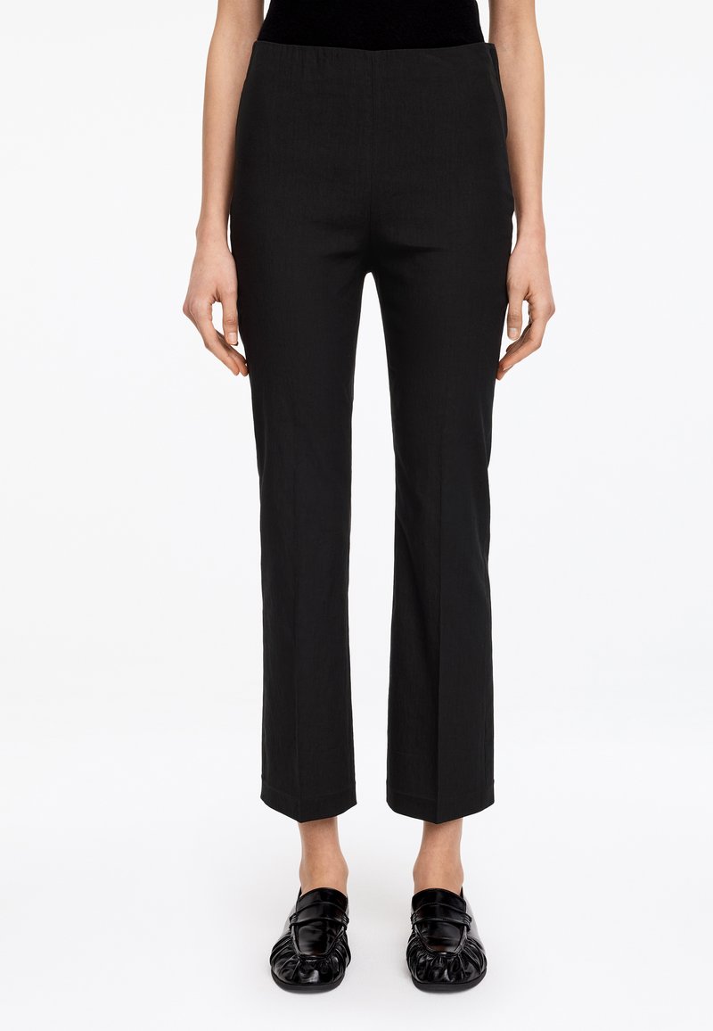 Femme portant un pantalon noir droit coupé à la cheville et des mocassins noirs sans lacets, debout devant un fond blanc.
