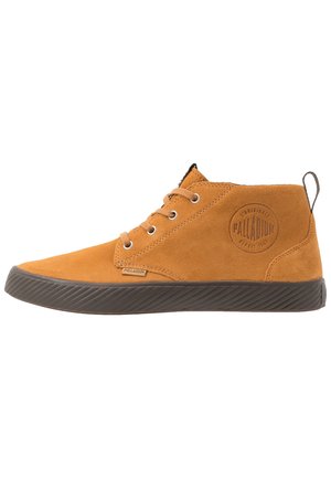 Baskets montantes en daim beige avec semelle en caoutchouc noire, lacets à l'avant, languette à l'arrière et logo rond Palladium sur le côté.