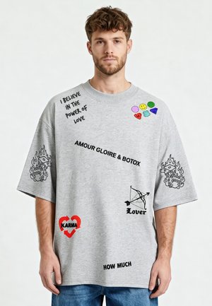 Jeune homme portant un t-shirt gris oversize avec des visages souriants colorés, des phrases en texte noir, des dés en flammes, un cœur rouge avec "KARMA" et un nœud étiqueté "Lover".