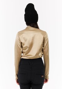 Langarm, gouden satijnen top met een gladde textuur, gecombineerd met zwarte broeken met een tailleband en twee achterzakken. Zwarte ribgebreide beanie.