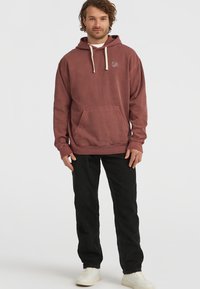 Bordeauxrode hoodie van katoen, met een frontzak, een capuchon met trekkoord en een klein logo op de borst. Gekoeld met zwarte broek en witte sneakers.