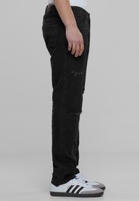 Mustat farkut slim fit -mallilla, joissa on reikiä ja kulutusta. Yhdistetty valkoisiin ja harmaisiin lenkkareihin. Sivuprofiili harmaata taustaa vasten.
