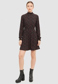 Vestido negro con un estampado floral en rojo, con cuello alto y mangas largas, cintura fruncida y falda acampanada. Combinado con botas negras hasta el tobillo.