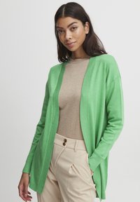 Cardigan vert en tissu doux, à ouverture sur le devant, avec des bords côtelés, superposé sur un haut beige et associé à un pantalon crème.