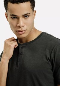 Ciemnozielona koszulka henley z krótkim rękawem, z trzyguzkowym zapięciem. Gładka tekstura, prążkowany kołnierzyk i dopasowany krój. Gładki materiał, bez wzorów.