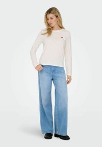 Langærmet, off-white bomulds t-shirt med et lille rødt hjerte-motiv på brystet, parret med brede blå denim jeans og sorte sko.