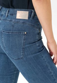 Donkerblauwe denim jeans hebben een klassiek vijf-pocket ontwerp, met een zichtbaar label "RAPHAELA" op de achterste tailleband.