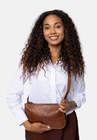 Sac en cuir marron porté en bandoulière, avec une texture lisse, une forme rectangulaire et une fermeture éclair, tenu par une personne en chemise blanche.