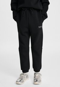 Person iført sorte joggingbukser med snøre og sølvfarvede sneakers, stående mod en ensfarvet baggrund.