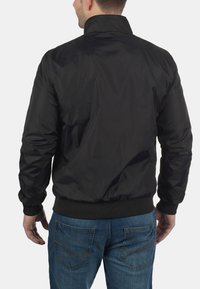 Černá bomber bunda s límcem, vyrobená z nylonového materiálu, s elastickými manžetami a pasem. Hladká textura s jednoduchým designem.
