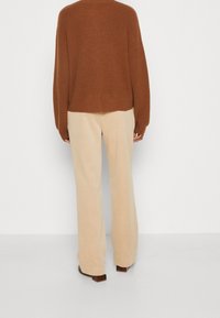 Brauner Strickpullover mit langen Ärmeln, kombiniert mit beigen, weit geschnittenen Hosen, die eine lockere Passform und sichtbare Textur aufweisen.