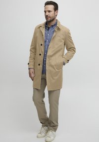 Manteau beige à designer simple boutonnage, avec de grands boutons noirs et des poches latérales. Porté sur une chemise bleu à boutons et un pantalon clair.