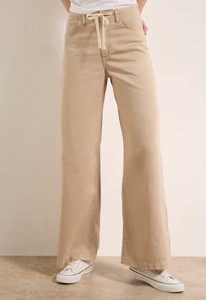 Beżowe spodnie o szerokich nogawkach z przednimi kieszeniami i sznurkiem w pasie, noszone z białymi trampkami na neutralnej podłodze i tle.