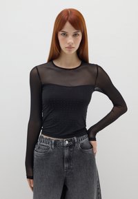 Schwarzes Langarmoberteil aus durchsichtiger Netzware mit gepunktetem Muster auf dem Oberteil und rundem Ausschnitt, kombiniert mit hoch taillierten grauen Jeans.