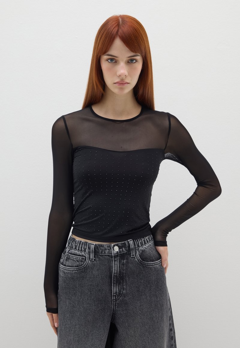 Schwarzes Langarmoberteil aus durchsichtiger Netzware mit gepunktetem Muster auf dem Oberteil und rundem Ausschnitt, kombiniert mit hoch taillierten grauen Jeans.