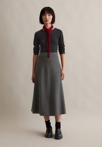 Fustă din lână gri cu o siluetă evazată, asortată cu un top negru slim cu mâneci lungi și o eșarfă roșie tricotată. Se observă pantofi negri și șosete.