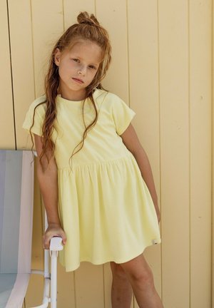 Jong meisje met lang haar in een gele jurk staat leunend op een witte en pastel gestreepte stoel tegen een bleekgele muur.