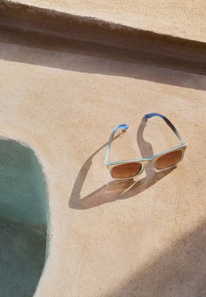 Occhiali da sole quadrati con montatura azzurro chiaro e lenti marroni, appoggiati su una superficie beige texturizzata. Ombra notevole castata dagli occhiali da sole.