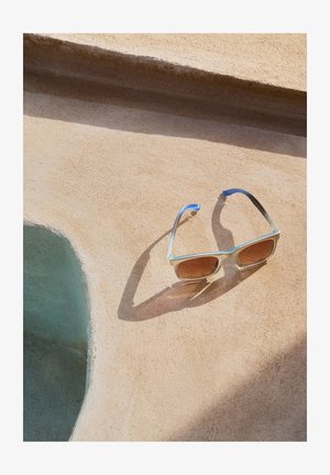 Gafas de sol cuadradas con montura azul claro y lentes marrones, descansando sobre una superficie beige texturizada. Sombra notable de las gafas de sol.