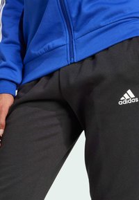 Blå zip-up jacka med svarta byxor, med Adidas-logotyp i vitt. Materialet verkar mjukt med en slät textur. Designen är enkel och atletisk.