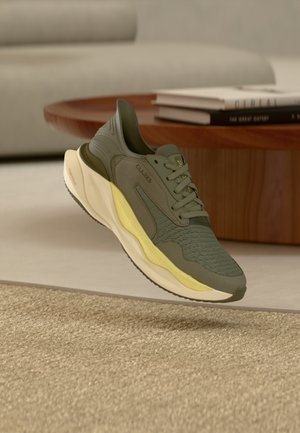 Olivgrön och gul Clarks-sneaker som svävar ovanför en beige mattbelagd golv, med ett träbord och böcker i bakgrunden.