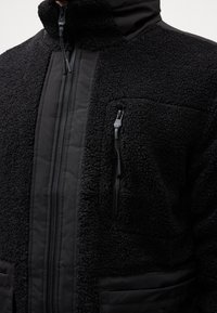 Veste polaire noire avec tissu texturé, fermeture éclair centrale, poche poitrine zippée et détails en tissu lisse au col et aux poches.