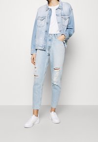 Giacca in denim azzurro chiaro con un lavaggio leggermente sbiadito, abbinata a jeans chiari strappati e sneakers bianche. I jeans presentano dettagli usurati.