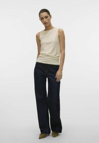 Beżowy bezrękawnik z detalem szwowym, zestawiony z ciemnoniebieskimi szerokimi spodniami i oliwkowymi szpiczastymi butami. Gładkie tekstury, casualowy styl.