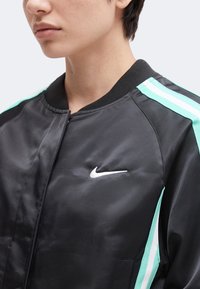 Γυναίκα που φοράει μαύρο μπουφάν Nike με τιρκουάζ και άσπρες ρίγες στους ώμους και τα μανίκια, δείχνοντας τον άνω κορμό και το λαιμό.