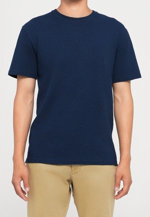 Homme portant un t-shirt uni bleu foncé à manches courtes et un pantalon marron clair, debout avec les bras détendus le long du corps.