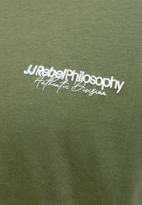 Tissu vert avec texte blanc indiquant "JJ Rebel Philosophy Authentic Division" dans deux styles de police différents.