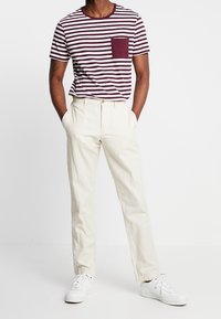 Camiseta de manga corta a rayas en burdeos y blanco con un bolsillo en el pecho, combinada con pantalones color crema y zapatillas blancas.