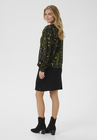 Frau in einer gemusterten grünen Bluse mit langen Ärmeln, schwarzem Rock und schwarzen Stiefeletten, die steht und über die Schulter schaut.