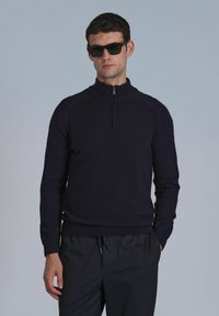 Marinefarbener Strickpullover mit hohem Kragen und kurzem Reißverschluss, getragen mit dunklen Hosen. Das Modell trägt auch dunkle Sonnenbrillen. Glatte Textur.