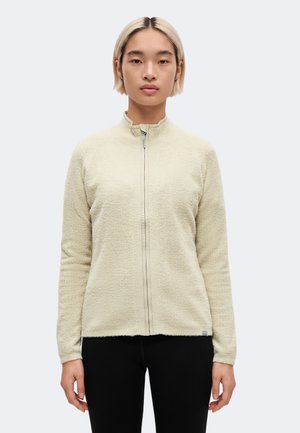 Une personne aux cheveux blonds courts est de face. Elle porte une veste polaire crème zippée et un pantalon noir, sur un fond clair uni.