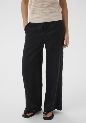 Nő laza fekete bőszárú nadrágban, bézs texturált ujjatlan felsőben és fekete, négyzetes orrú, lábujjközi pántos szandálban.