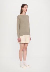 Kaki strikkegenser med ribbestrikket kant, kombinert med beige shorts med horisontal stripe, og hvite sokker med spisse hæler.
